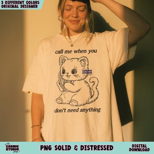 May include: Cream-colored t-shirt featuring a black and white illustration of a cartoon cat holding a phone. The text "call me when you don't need anything" is above and below the cat. The shirt also has the text "Cosmic Studio".