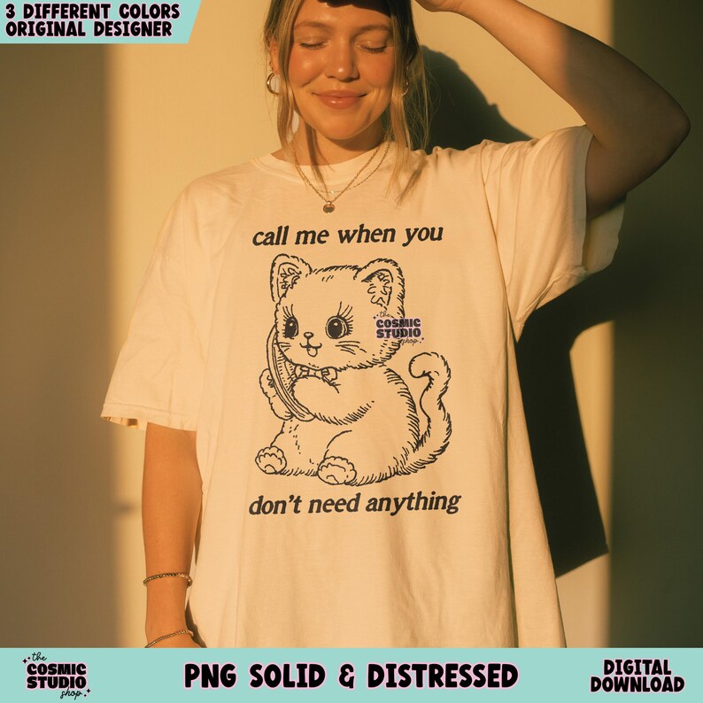 May include: Cream-colored t-shirt featuring a black and white illustration of a cartoon cat holding a phone. The text "call me when you don't need anything" is above and below the cat. The shirt also has the text "Cosmic Studio".