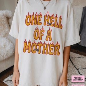 May include: Cream-colored t-shirt with the text "ONE HELL OF A MOTHER" in yellow and orange flame-style lettering. The shirt features a crew neck and short sleeves. The Cosmic Studio shop logo is visible.