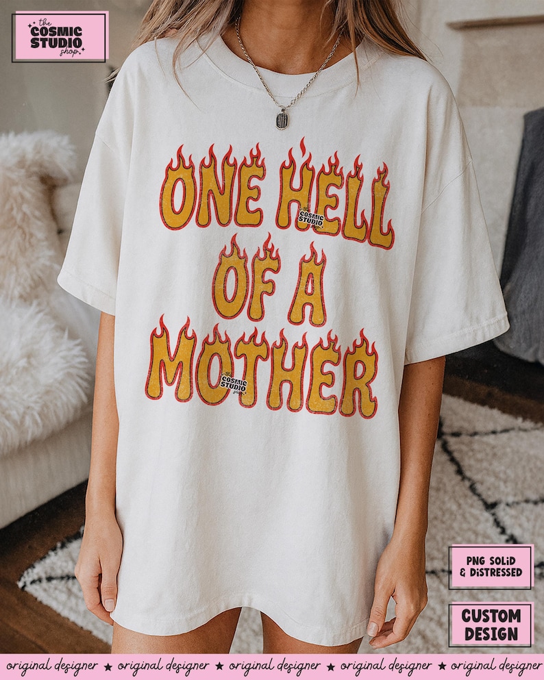 May include: Cream-colored t-shirt with the text "ONE HELL OF A MOTHER" in yellow and orange flame-style lettering. The shirt features a crew neck and short sleeves. The Cosmic Studio shop logo is visible.