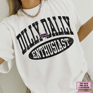 K&ouml;nnte beinhalten: Ein wei&szlig;es kurz&auml;rmeliges T-Shirt mit schwarzem, abgenutztem Text, der DILLY DALLY ENTHUSIAST lautet. Die Worte DILLY DALLY sind in einer gro&szlig;en, gebogenen Schriftart gehalten, w&auml;hrend ENTHUSIAST in einem schwarzen Oval steht. Das T-Shirt hat einen Rundhalsausschnitt.