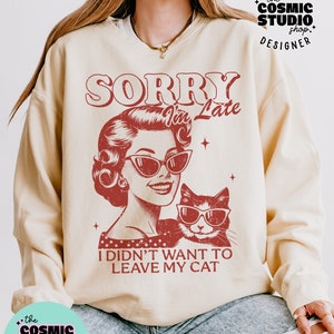 May include: Cream-colored sweatshirt with a distressed red graphic. The graphic features a vintage illustration of a woman and a cat, both wearing sunglasses, with the text "SORRY I'm Late, I didn't want to leave my cat."
