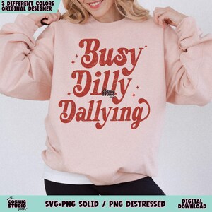 May include: A light pink sweatshirt with the words "Busy Dilly Dallying" in a retro red font. The sweatshirt has a crew neck and long sleeves. The Cosmic Studio shop logo is at the bottom.