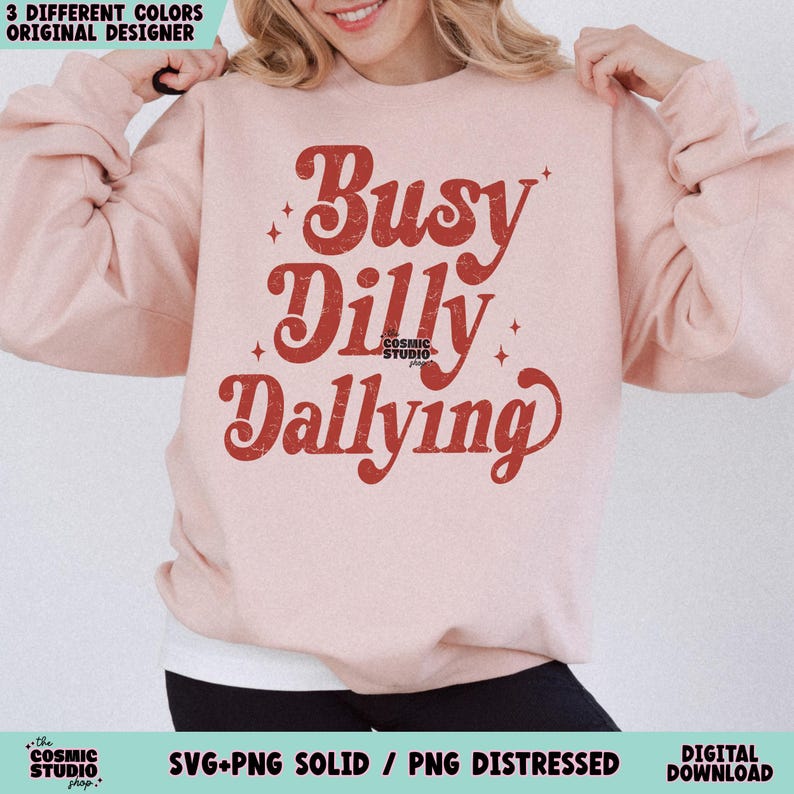 May include: A light pink sweatshirt with the words "Busy Dilly Dallying" in a retro red font. The sweatshirt has a crew neck and long sleeves. The Cosmic Studio shop logo is at the bottom.