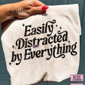 May include: A cream-coloured t-shirt with the text "Easily Distracted by Everything" in a black, retro font. The shirt also features small star graphics. The Cosmic Studio shop logo is in the upper left corner.