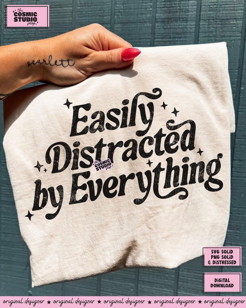 May include: A cream-coloured t-shirt with the text "Easily Distracted by Everything" in a black, retro font. The shirt also features small star graphics. The Cosmic Studio shop logo is in the upper left corner.