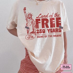 以下が含まれることがあります： クリーム色のTシャツ。赤色の自由の女神のグラフィックと「Land of the Free, 250 Years, Since 1776, Home of the Brave」の文字。デジタルダウンロードです。