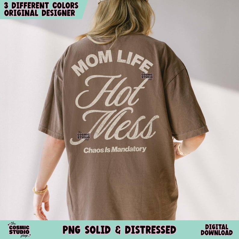 May include: A brown t-shirt with the text "MOM LIFE Hot Mess Chaos Is Mandatory" in a distressed white font. The shirt has short sleeves and a relaxed fit. The Cosmic Studio logo is also visible.