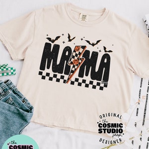 以下が含まれることがあります： ベージュのTシャツに、黒いグラフィックで「ママ」と書いてあり、稲妻とコウモリが描かれています。グラフィックは市松模様の背景になっています。「ORIGINAL the COSMIC STUDIO shop. DESIGNER」というテキストがグラフィックの下に印刷されています。