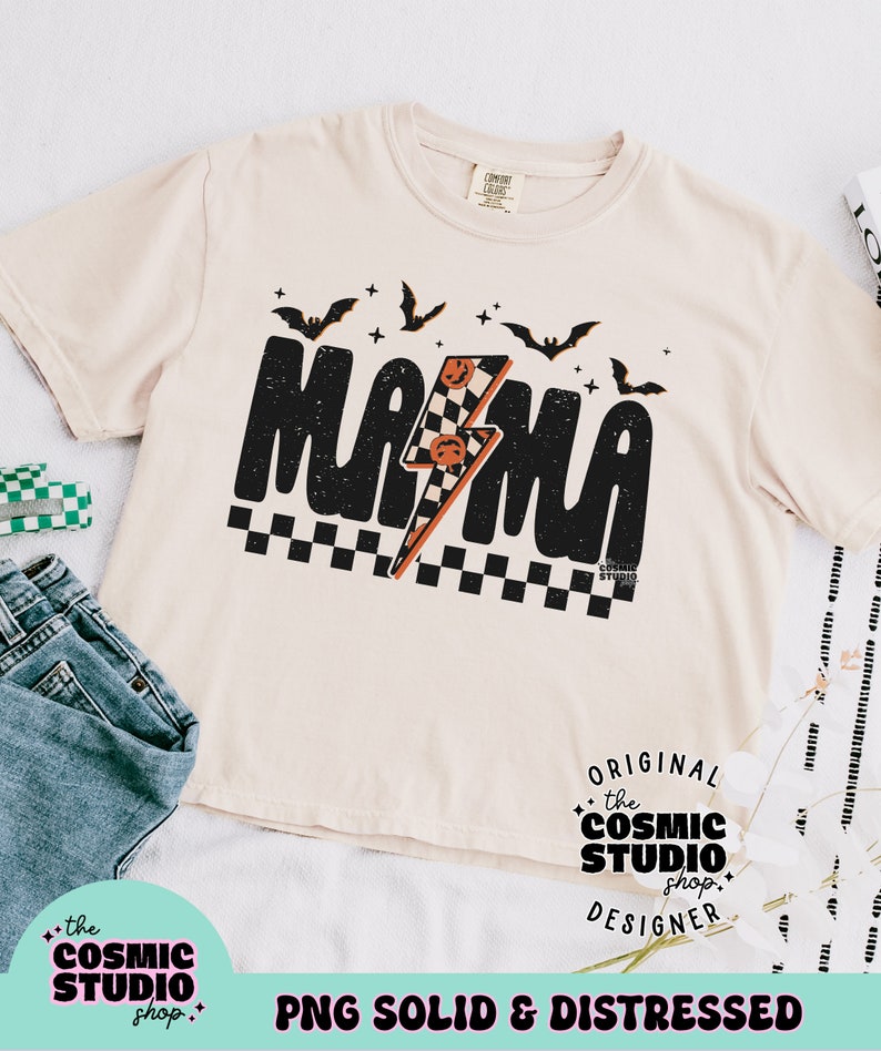 以下が含まれることがあります： ベージュのTシャツに、黒いグラフィックで「ママ」と書いてあり、稲妻とコウモリが描かれています。グラフィックは市松模様の背景になっています。「ORIGINAL the COSMIC STUDIO shop. DESIGNER」というテキストがグラフィックの下に印刷されています。