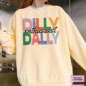 May include: A light yellow sweatshirt with the text "DILLY enthusiast DALLY" in colorful, distressed lettering. The design includes a star and the words "the COSMIC STUDIO shop". The sweatshirt is a digital download.