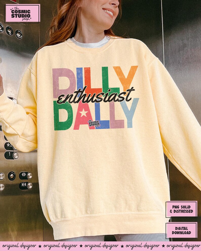 May include: A light yellow sweatshirt with the text "DILLY enthusiast DALLY" in colorful, distressed lettering. The design includes a star and the words "the COSMIC STUDIO shop". The sweatshirt is a digital download.