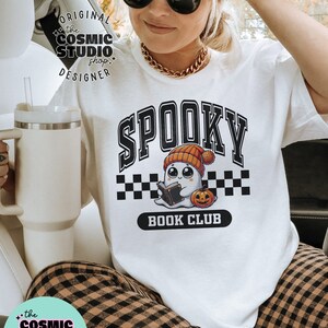 May include: White t-shirt featuring a black and white graphic of a ghost wearing a hat and holding a book. The graphic reads "Spooky Book Club".