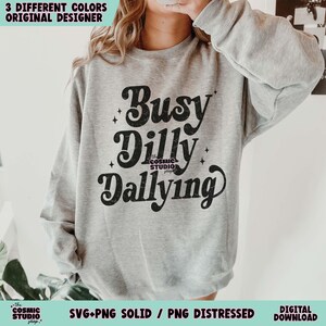 May include: A light gray sweatshirt with the words "Busy Dilly Dallying" in a black, stylized font. The text is the focal point, with the words arranged in a playful, stacked design. The Cosmic Studio shop logo is at the bottom.