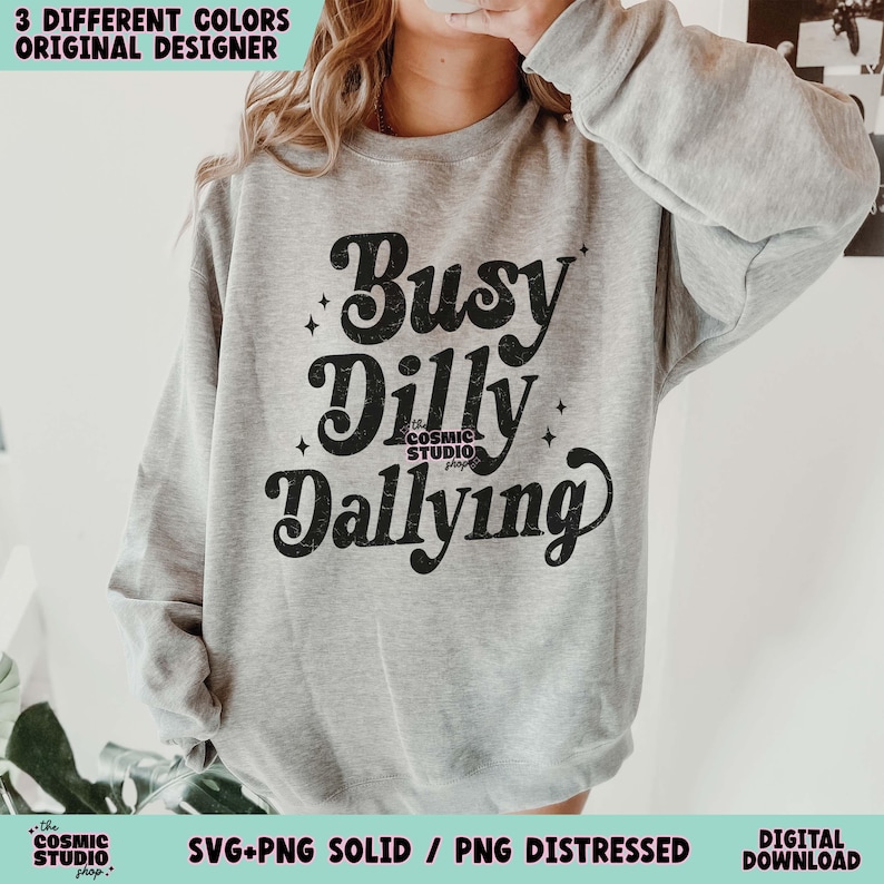 May include: A light gray sweatshirt with the words "Busy Dilly Dallying" in a black, stylized font. The text is the focal point, with the words arranged in a playful, stacked design. The Cosmic Studio shop logo is at the bottom.