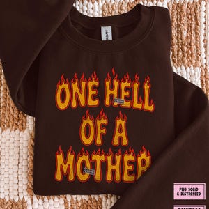 May include: A dark brown sweatshirt with the text "ONE HELL OF A MOTHER" in orange and red flame-style lettering. The sweatshirt has a crew neck and long sleeves. The Cosmic Studio logo is visible.