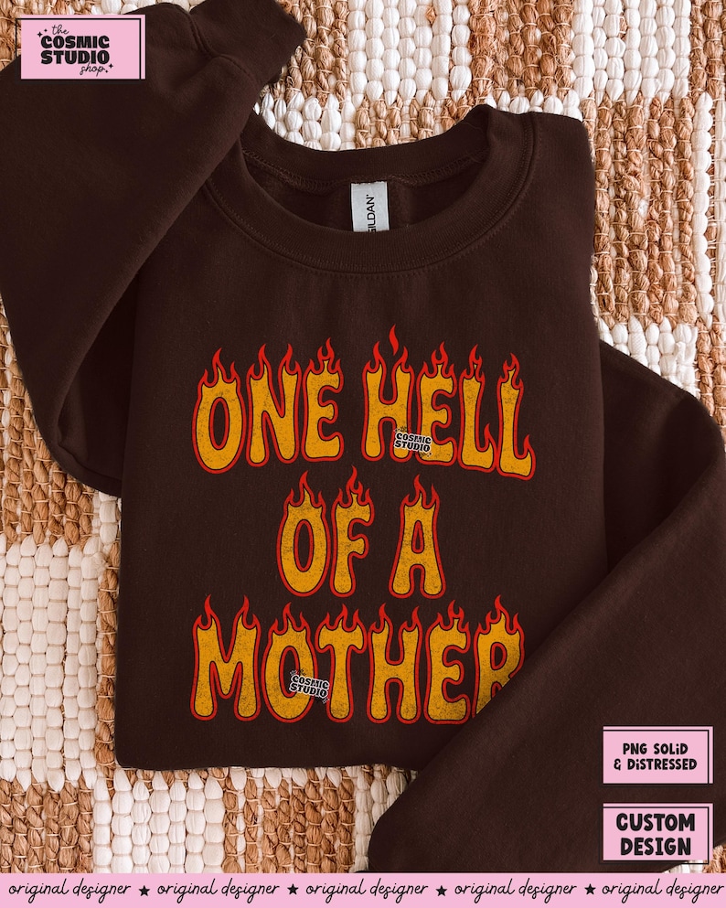 May include: A dark brown sweatshirt with the text "ONE HELL OF A MOTHER" in orange and red flame-style lettering. The sweatshirt has a crew neck and long sleeves. The Cosmic Studio logo is visible.