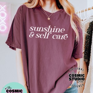May include: A maroon t-shirt with the text "sunshine & self care" in white. The t-shirt is being worn by a person. The image also includes a logo for "the cosmic studio shop" in black and white.