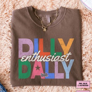 May include: A brown crew neck t-shirt with the words "DILLY enthusiast DALLY" in colorful, distressed lettering. A gold chain necklace with a pendant rests on the shirt. The Cosmic Studio shop logo is visible.