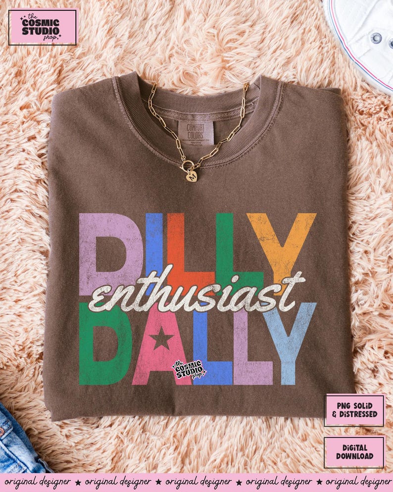 May include: A brown crew neck t-shirt with the words "DILLY enthusiast DALLY" in colorful, distressed lettering. A gold chain necklace with a pendant rests on the shirt. The Cosmic Studio shop logo is visible.