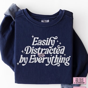 May include: A navy blue sweatshirt featuring the white text "Easily Distracted by Everything" with star details. The Cosmic Studio shop logo is also visible. The sleeves have a grey accent.