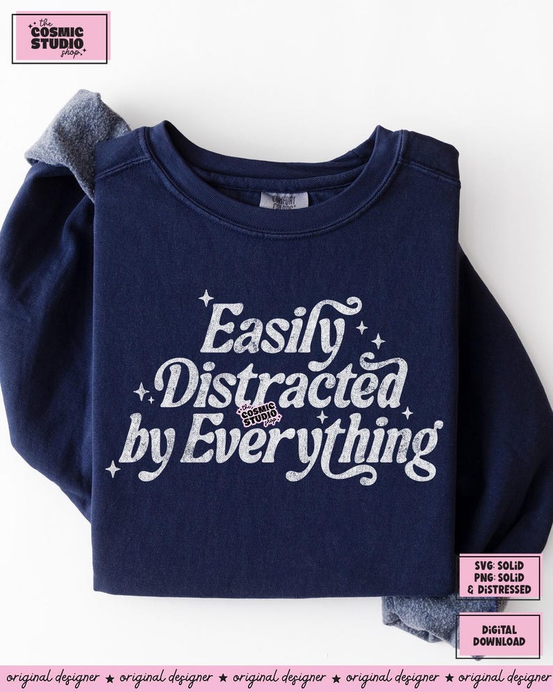 May include: A navy blue sweatshirt featuring the white text "Easily Distracted by Everything" with star details. The Cosmic Studio shop logo is also visible. The sleeves have a grey accent.