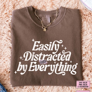 May include: A brown t-shirt with the slogan "Easily Distracted by Everything" in white. The shirt is folded and has a crew neck. A gold chain necklace with a pendant rests on the shirt.