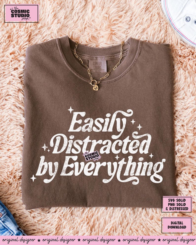 May include: A brown t-shirt with the slogan "Easily Distracted by Everything" in white. The shirt is folded and has a crew neck. A gold chain necklace with a pendant rests on the shirt.