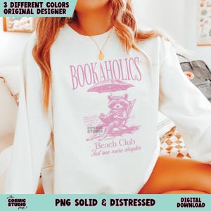 May include: A white sweatshirt with a pink graphic design featuring a raccoon reading a book under an umbrella. The text "BOOKAHOLICS" is at the top of the design, and the text "Beach Club Just one more chapter" is at the bottom.