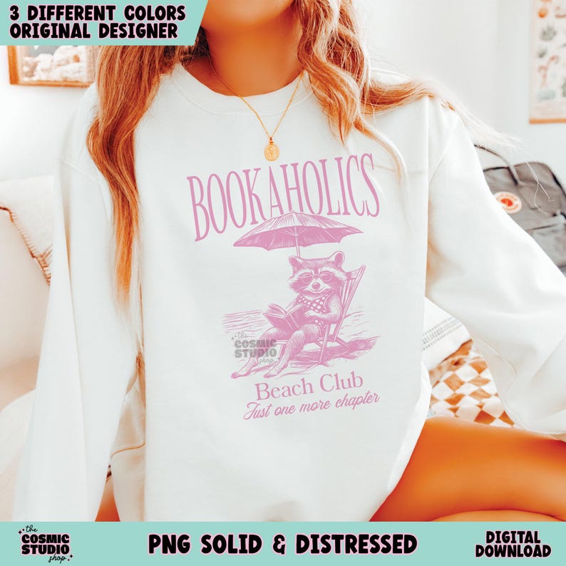 May include: A white sweatshirt with a pink graphic design featuring a raccoon reading a book under an umbrella. The text "BOOKAHOLICS" is at the top of the design, and the text "Beach Club Just one more chapter" is at the bottom.