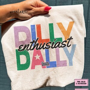 May include: Cream-colored t-shirt with the words "DILLY enthusiast DALLY" in colorful block letters. The design includes a star and the text "the COSMIC STUDIO shop". The shirt is slightly crumpled.