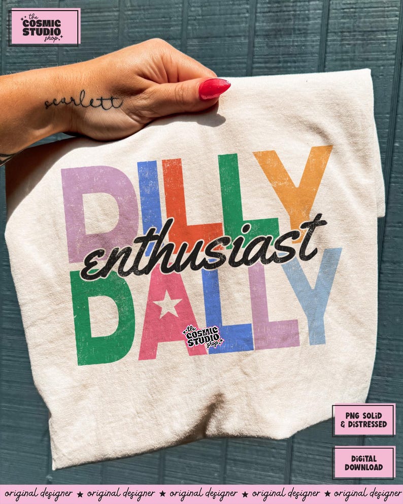 May include: Cream-colored t-shirt with the words "DILLY enthusiast DALLY" in colorful block letters. The design includes a star and the text "the COSMIC STUDIO shop". The shirt is slightly crumpled.
