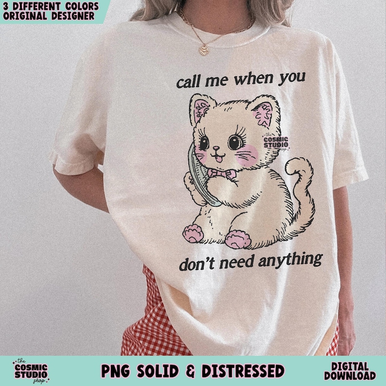 May include: Cream-colored t-shirt featuring a cartoon cat holding a phone, with the text "call me when you don't need anything." The cat has pink accents and a bow. The shirt also has the text "the COSMIC STUDIO shop."