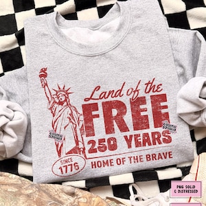 以下が含まれることがあります： 自由の女神と「Land of the FREE 250 YEARS HOME OF THE BRAVE SINCE 1776」の文字がプリントされた、グレーのスウェットシャツ。デザインには、Cosmic Studio shopのロゴも含まれています。