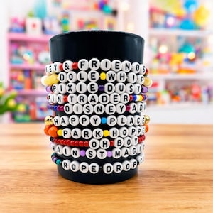 May include: A black cylindrical container adorned with multiple beaded bracelets. The bracelets feature white letter beads spelling out phrases like "KEY & FRIENDS", "DOLE WHIP", and "ROPE DROP", interspersed with colorful beads.