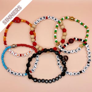 May include: A collection of five beaded bracelets featuring various colored beads, including red, orange, yellow, blue, green, and black. The bracelets incorporate letter beads spelling phrases like CLUB JUKE, DELTA DEMON, SMOKE, and DANCE WITH THE DEVIL.