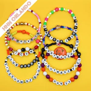 May include: A collection of ten colorful beaded bracelets featuring white letter beads spelling out phrases including HHN ORLANDO, DOWNTOWN CLOWN ARES, FORTNITE MARES, NIGHTMARE FUEL, CLUB HORROR, SIDESHOW OF DECAY, and ICON. The bracelets use various plastic beads.