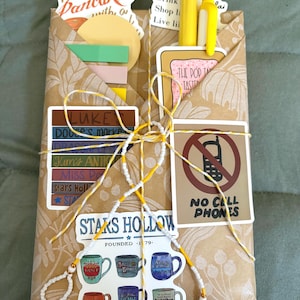 May include: A brown paper envelope decorated with various stickers including text reading I'M GOING TO HAVE PANCAKES, STARS HOLLOW, and NO CELL PHONES. The envelope is tied with yellow and white string and contains yellow pens and colorful sticky notes.