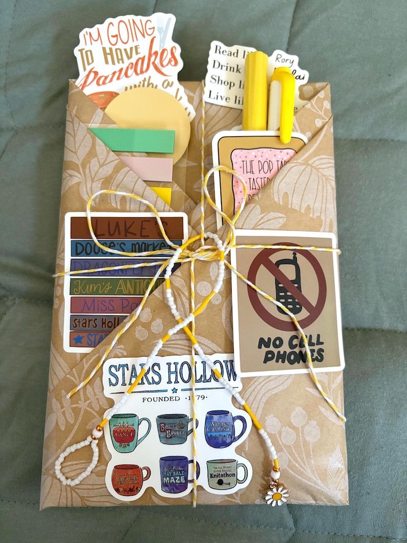 May include: A brown paper envelope decorated with various stickers including text reading I'M GOING TO HAVE PANCAKES, STARS HOLLOW, and NO CELL PHONES. The envelope is tied with yellow and white string and contains yellow pens and colorful sticky notes.