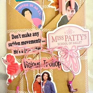 May include: A tan plaid paper envelope decorated with various stickers including Miss Patty's School of Ballet, a pink poodle, and characters from Gilmore Girls. The text reads Don't make any sudden movements, Coffee in an L.V., and Vicious Trollop. It is tied with pink and white twine featuring a small boot charm.