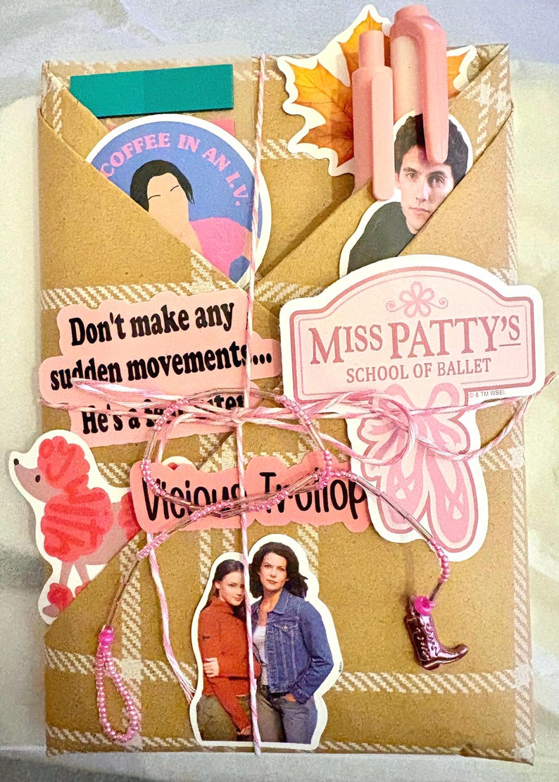 May include: A tan plaid paper envelope decorated with various stickers including Miss Patty's School of Ballet, a pink poodle, and characters from Gilmore Girls. The text reads Don't make any sudden movements, Coffee in an L.V., and Vicious Trollop. It is tied with pink and white twine featuring a small boot charm.