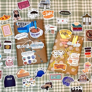 May include: Two brown paper-wrapped packages tied with string, surrounded by numerous colorful stickers featuring various illustrations, icons, and text related to the television show Gilmore Girls, including coffee cups, books, and signs.