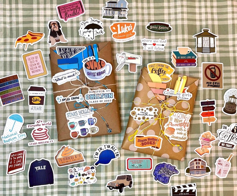 May include: Two brown paper-wrapped packages tied with string, surrounded by numerous colorful stickers featuring various illustrations, icons, and text related to the television show Gilmore Girls, including coffee cups, books, and signs.