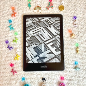 Peut inclure: Une liseuse Kindle noire et blanche entourée de marque-pages en forme d'animaux en ballon de couleurs. Les marque-pages sont en plastique et présentent une variété de motifs, notamment un arc-en-ciel, un éclair, un patin à roulettes, une boule disco et une pièce d'or. Les marque-pages sont disposés en cercle autour du Kindle.