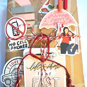 May include: A rectangular package wrapped in brown paper with a white plaid pattern, tied with red and white baker's twine. It is decorated with various stickers including a no cell phones sign, a pink umbrella, and text reading life's short talk fast.
