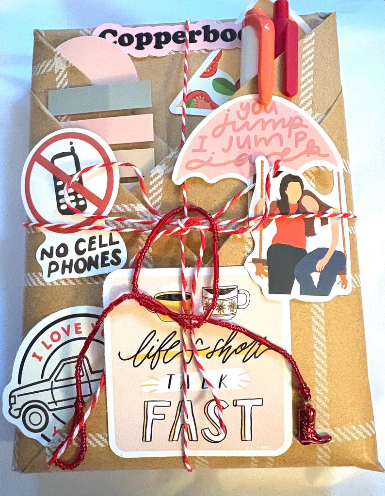 May include: A rectangular package wrapped in brown paper with a white plaid pattern, tied with red and white baker's twine. It is decorated with various stickers including a no cell phones sign, a pink umbrella, and text reading life's short talk fast.