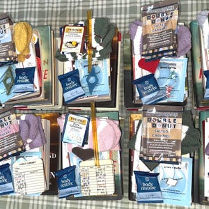 May include: A collection of bath and body products arranged on a table. The products include bath bombs, body scrubs, and bath salts. The bath bombs are in the shape of donuts and are labeled with different flavors, such as "Double Donut Chocolate Raspberry" and "Double Donut Salted Caramel". The body scrubs are in small jars and are labeled with different scents, such as "Peppermint Patty" and "Lemon & Ginger". The bath salts are in small bags and are labeled with different scents, such as "Pomegranate & Raspberry".
