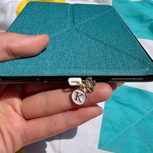 May include: Teal tablet case with a gold charm. The charm features a white circular pendant with the letter "K" and a gold flower. The case is held in a hand.