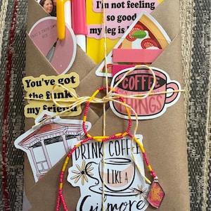 May include: A brown paper-wrapped package decorated with various stickers including a pizza slice, a gazebo, and coffee-themed text. It features a pink pen, a yellow pen, and a yellow and red beaded string tied around the center with a small charm.