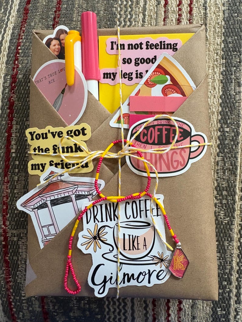 May include: A brown paper-wrapped package decorated with various stickers including a pizza slice, a gazebo, and coffee-themed text. It features a pink pen, a yellow pen, and a yellow and red beaded string tied around the center with a small charm.